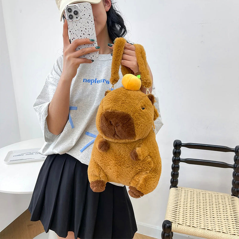 Close-up of a plush capybara backpack being held by its fuzzy straps, showcasing cute orange fruit on top.