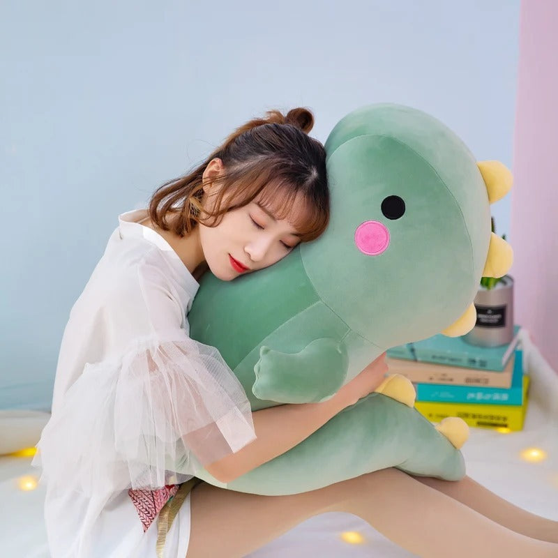 Large green dinosaur plush from the Kawaii collection, a soft and squishable giant dino stuffed animal being hugged by a woman to show its jumbo size. This cute plushie features yellow back spikes, rosy pink cheeks, and a round body, making it the perfect huggable companion for kawaii room decor.