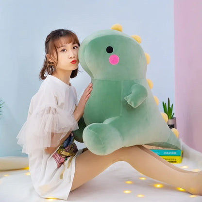 Large green dinosaur plushie from the Kawaii collection, a soft and squishable giant dino stuffed animal for bedroom decor. Features include yellow back spikes, pink rosy cheeks, and a huggable round body, shown held by a person to demonstrate its jumbo size