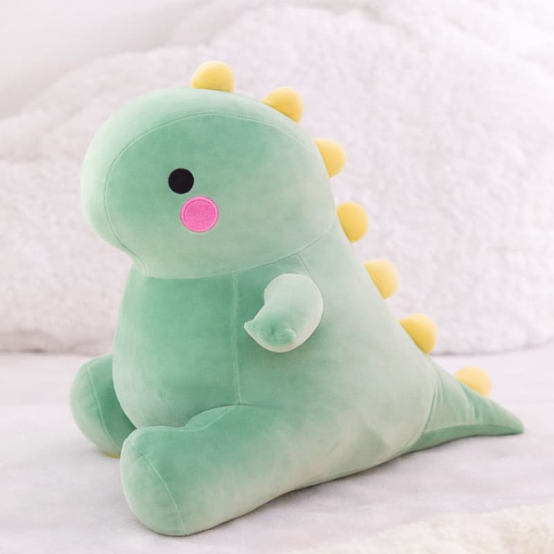 Octoplush Kawaii collection green dinosaur plushie, a soft and squishable dino stuffed animal sitting on a white bed. This cute Octoplush.store toy features a round pastel green body, rosy pink cheeks, and yellow triangular spikes, perfect for aesthetic room decor and cozy gifts.