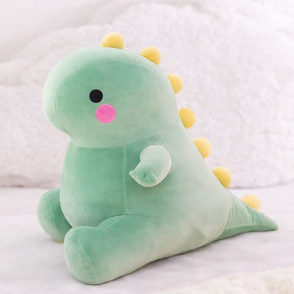 Octoplush Kawaii collection green dinosaur plushie, a soft and squishable dino stuffed animal sitting on a white bed. This cute Octoplush.store toy features a round pastel green body, rosy pink cheeks, and yellow triangular spikes, perfect for aesthetic room decor and cozy gifts.