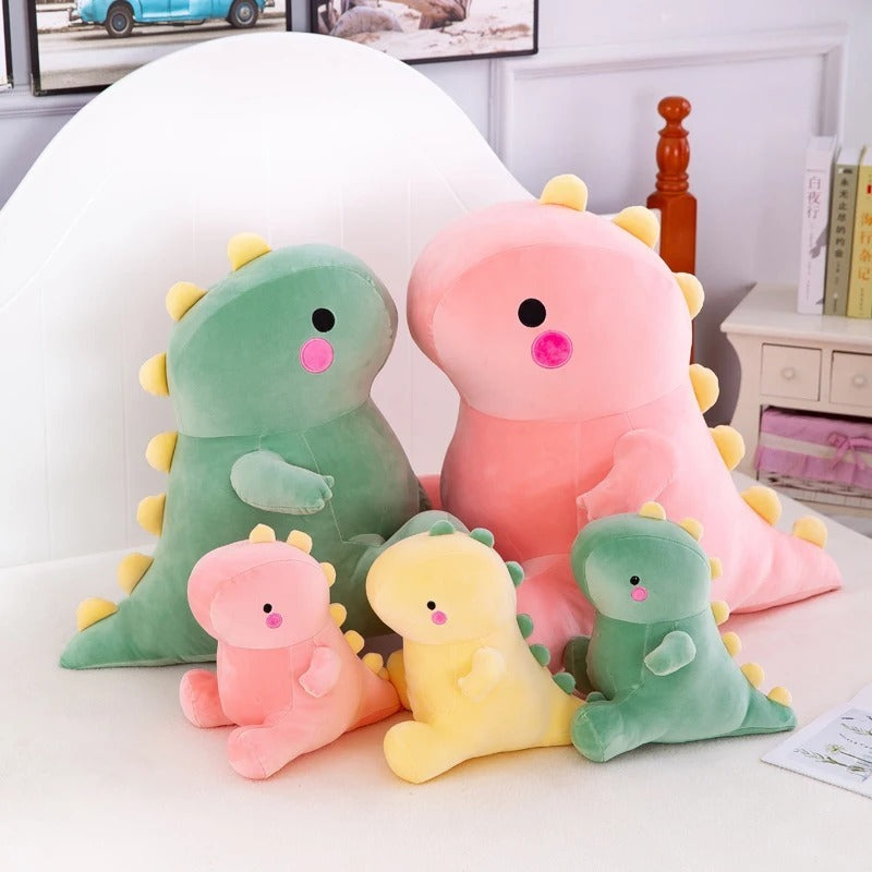 The Octoplush Kawaii collection featuring a family of soft and squishable dinosaur plushies in pastel pink, green, and yellow. This assortment of cute dino stuffed animals includes various sizes from small to large, all with signature yellow spikes and rosy cheeks, perfect for aesthetic bedroom decor and gifts.