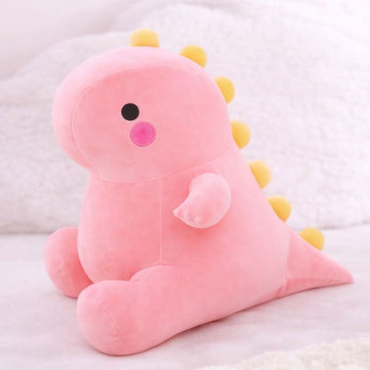 Octoplush Kawaii collection pink dinosaur plushie, a soft and squishable pastel pink dino stuffed animal sitting on a white bed. This cute Octoplush.store toy features yellow back spikes and bright pink rosy cheeks, making it an essential piece for aesthetic room decor and the perfect gift for girls.