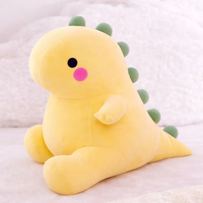 Octoplush Kawaii collection yellow dinosaur plushie, a soft and squishable pastel dino stuffed animal sitting on a white surface. This cute Octoplush.store toy features green back spikes and rosy pink cheeks, making it a perfect gift for aesthetic room decor and plushie collectors.